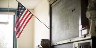 An American Flag hangs in the front of a classroom near a chalk board.