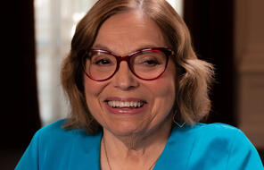 A headshot of Judy Heumann, a white woman with shoulder-length brown hair wearing red glasses, a blue v-neck shirt, and a gold necklace. She is smiling warmly.